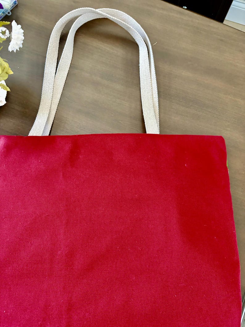 Op de afbeelding: Een effen rode draagtas met twee stevige, lichtbeige geweven stoffen hengsels die aan de bovenrand zijn bevestigd. De tas heeft een rechthoekige vorm met een plat, effen voorvlak gemaakt van duurzaam, ondoorzichtig textielmateriaal.