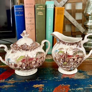 May include: A vintage cream and sugar set with a detailed brown and white transferware design. The set includes a lidded sugar bowl and a creamer, both featuring a scenic illustration. Books are in the background.