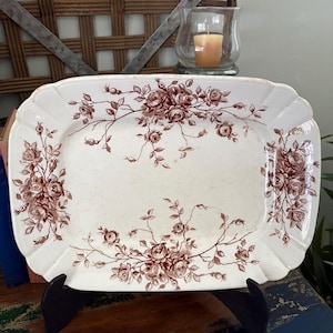 May include: A rectangular, antique white serving platter with a brown floral rose pattern. The platter has a scalloped edge and is displayed on a black stand. A candle in a glass holder and a woven basket are in the background.