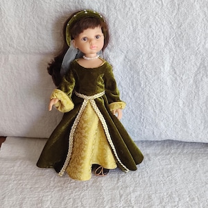 May include: A doll with brown hair wearing a dark green velvet dress with long sleeves, a gold patterned underskirt, and a braided gold trim. The doll features a pearl necklace, a matching green velvet headband with small pearls, and a sheer white veil.