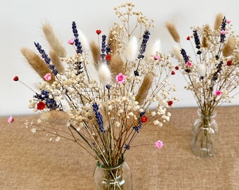 May include: Two glass vases filled with dried flowers. The flowers are a mix of white, brown, pink, and purple. The vases are sitting on a brown burlap surface.