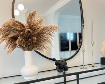 May include: A black metal console table with glass shelves, featuring a white vase with dried pampas grass, a black knotted sculpture, two gold candle holders with clear candles, and a white ceramic vase with gold polka dots.