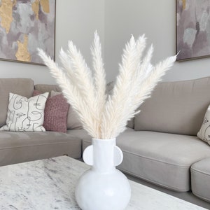 May include: White pampas grass in a white ceramic vase with two round handles. The vase is sitting on a white marble table in front of a light grey couch.