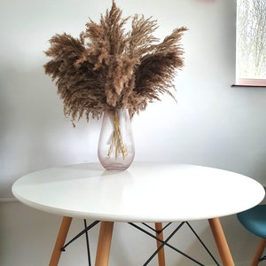 May include: A white round table with a wooden base and black metal legs. A vase with dried pampas grass is on the table.