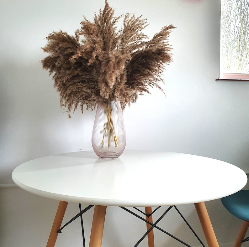 May include: A white round table with a wooden base and black metal legs. A vase with dried pampas grass is on the table.