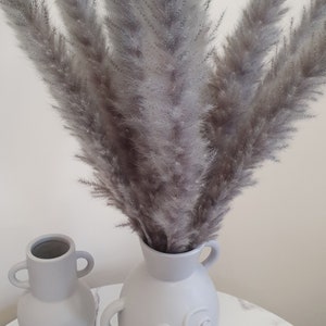 May include: Two gray ceramic vases with a face design. The vase on the right has a bouquet of dried gray pampas grass.