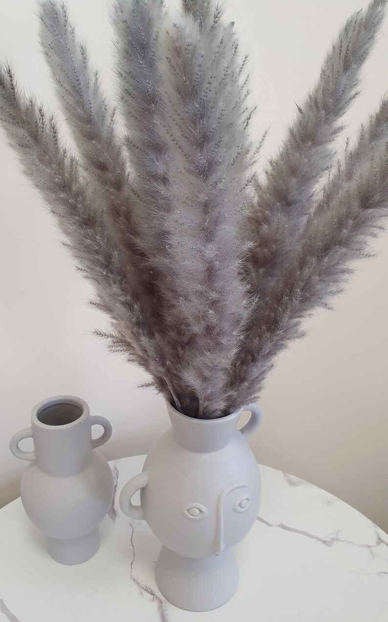 May include: Two gray ceramic vases with a face design. The vase on the right has a bouquet of dried gray pampas grass.