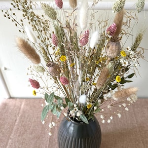 May include: A dried flower arrangement in a dark gray vase. The bouquet features a variety of dried grasses, flowers, and seed pods in shades of beige, brown, pink, and white.