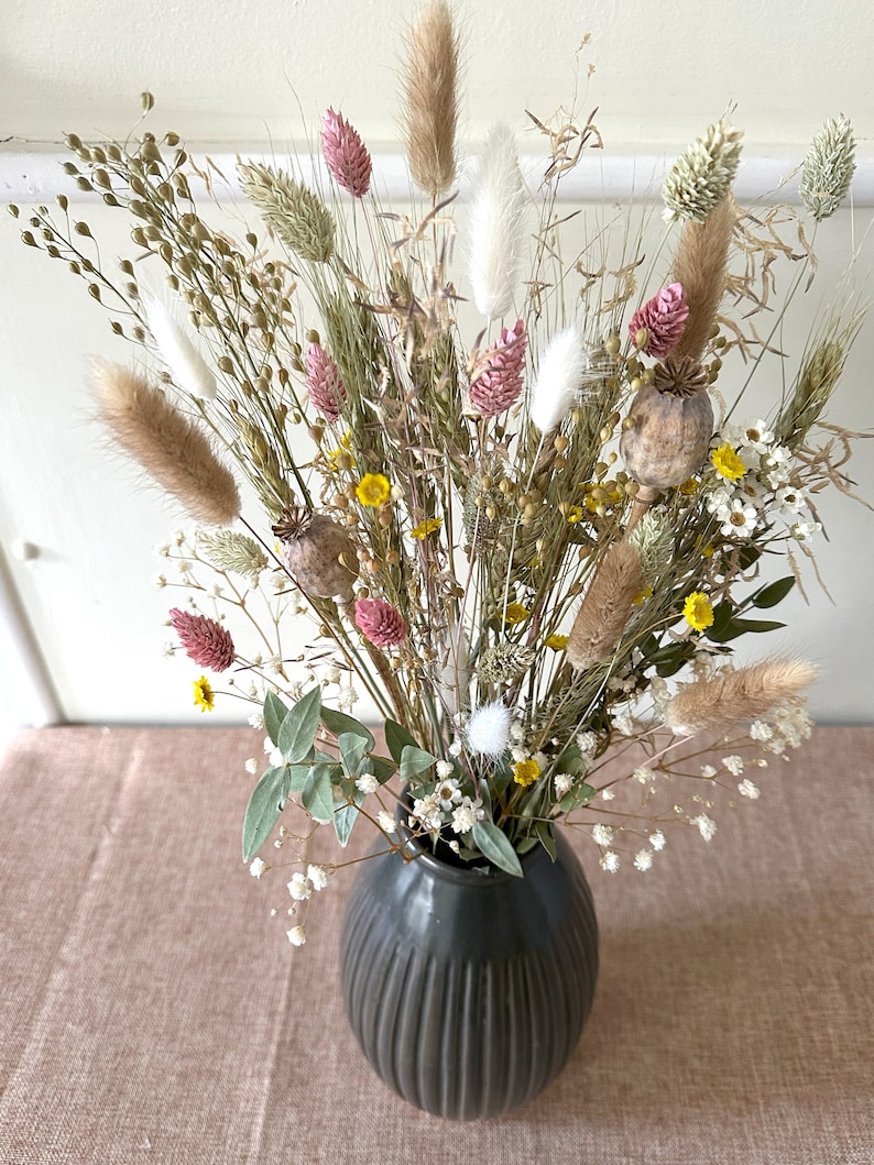 May include: A dried flower arrangement in a dark gray vase. The bouquet features a variety of dried grasses, flowers, and seed pods in shades of beige, brown, pink, and white.