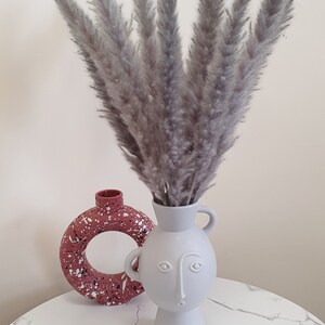 May include: A white ceramic vase with a face design holds a bouquet of dried pampas grass. The vase is sitting on a white marble surface next to a pink and white speckled vase.