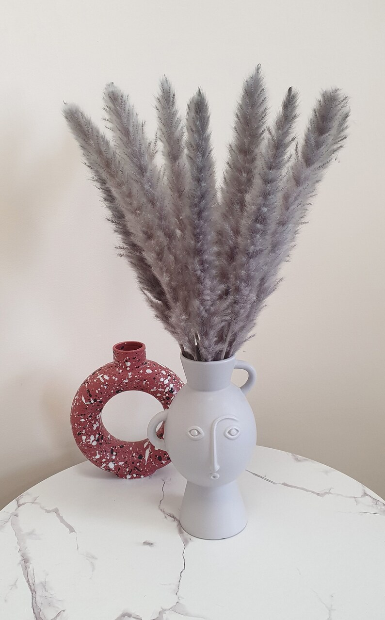 May include: A white ceramic vase with a face design holds a bouquet of dried pampas grass. The vase is sitting on a white marble surface next to a pink and white speckled vase.