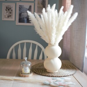 May include: A white vase with a bulbous shape holds a bouquet of dried pampas grass. The vase is sitting on a woven placemat on a wooden table. A small glass cloche with a candle inside sits on the table in front of the vase.
