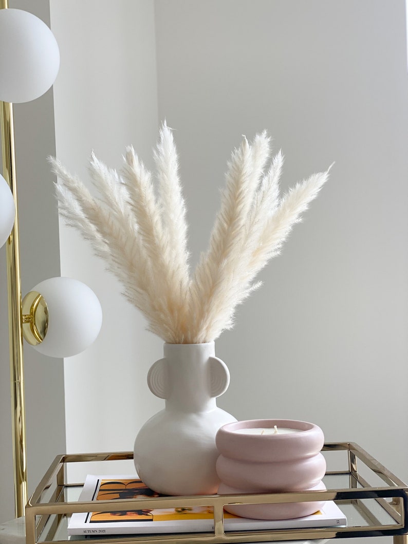 May include: A white ceramic vase with two handles on the sides, filled with dried pampas grass. A pink candle in a textured container sits on a gold tray with a magazine on top.