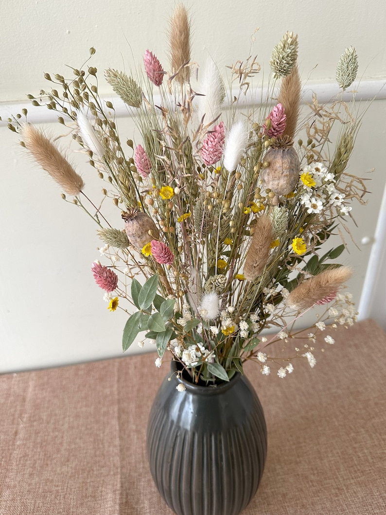May include: A dried flower bouquet in a dark gray vase. The bouquet features a variety of dried grasses, flowers, and seed pods in shades of pink, white, yellow, and brown.