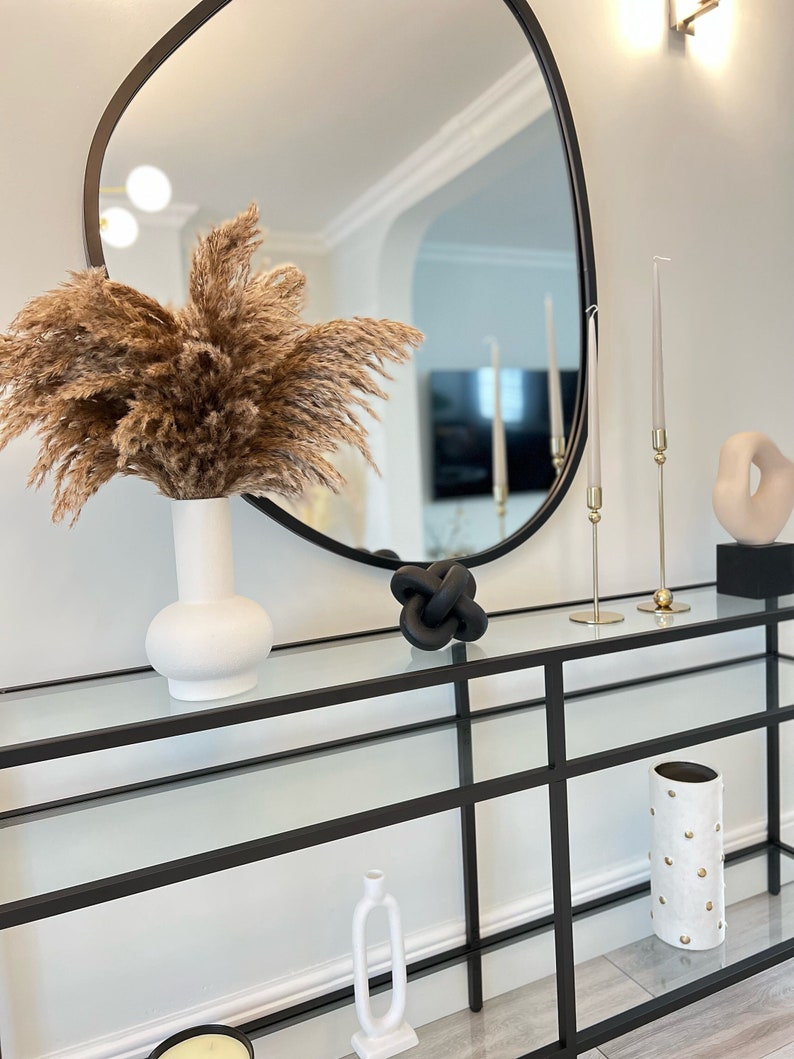 May include: A black metal console table with glass shelves, featuring a white vase with dried pampas grass, a black knotted sculpture, two gold candle holders with clear candles, and a white ceramic vase with gold polka dots.
