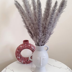 May include: A gray ceramic vase with a face design holds a bouquet of dried pampas grass. The vase is sitting on a white marble table next to a red speckled ceramic vase.
