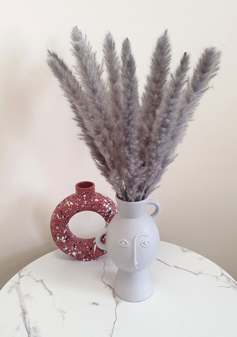 May include: A gray ceramic vase with a face design holds a bouquet of dried pampas grass. The vase is sitting on a white marble table next to a red speckled ceramic vase.