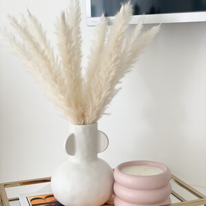 May include: A white ceramic vase with two handles, filled with dried pampas grass, sits on a gold metal tray with a pink candle in a textured container. A magazine is visible underneath the tray.
