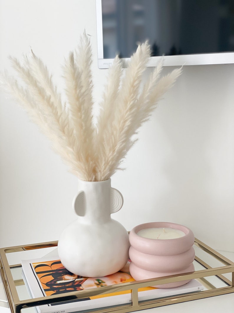 May include: A white ceramic vase with two handles, filled with dried pampas grass, sits on a gold metal tray with a pink candle in a textured container. A magazine is visible underneath the tray.