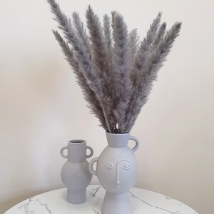 May include: Two gray ceramic vases with a face design. The larger vase holds a bouquet of dried gray pampas grass.