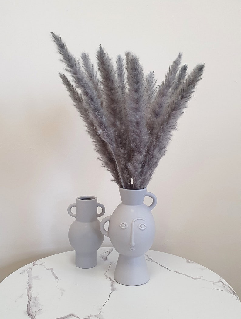 May include: Two gray ceramic vases with a face design. The larger vase holds a bouquet of dried gray pampas grass.