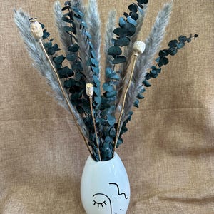 May include: A white ceramic vase with a stylized face design holds a bouquet of dried pampas grass and eucalyptus.