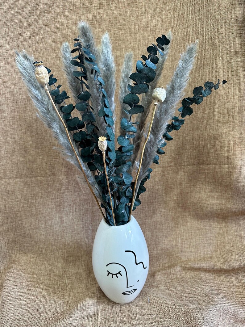 May include: A white ceramic vase with a stylized face design holds a bouquet of dried pampas grass and eucalyptus.