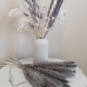 May include: A bouquet of dried pampas grass in a white vase. The bouquet is a mix of white and gray pampas grass. The vase is on a white marble table.