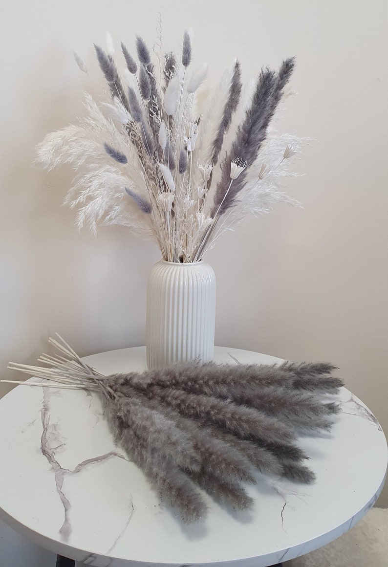 May include: A bouquet of dried pampas grass in a white vase. The bouquet is a mix of white and gray pampas grass. The vase is on a white marble table.