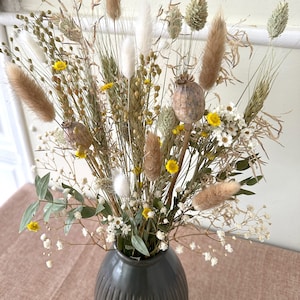 May include: A dried flower arrangement in a dark gray vase. The bouquet features a variety of dried grasses, fluffy white and brown pampas grass, small yellow flowers, and white seed pods.