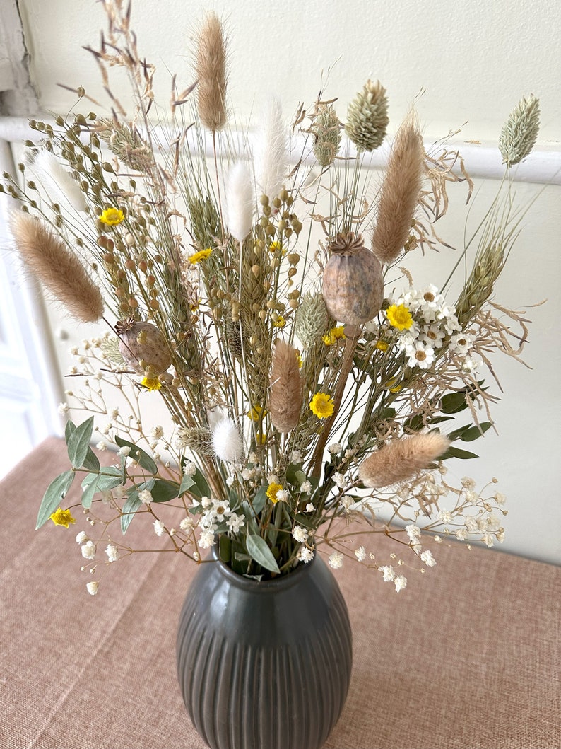May include: A dried flower arrangement in a dark gray vase. The bouquet features a variety of dried grasses, fluffy white and brown pampas grass, small yellow flowers, and white seed pods.