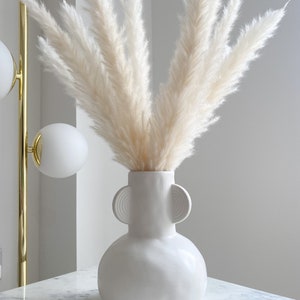 May include: White pampas grass in a white ceramic vase with two round handles. The vase has a textured surface and is sitting on a white marble surface.