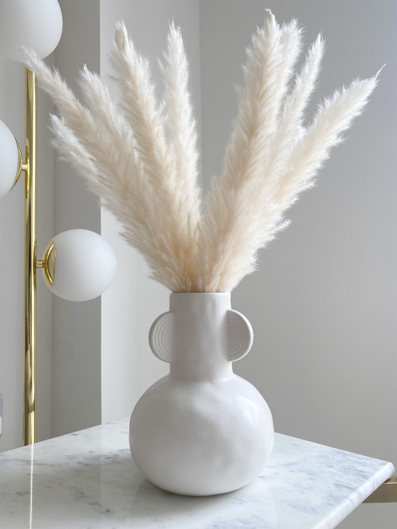 May include: White pampas grass in a white ceramic vase with two round handles. The vase has a textured surface and is sitting on a white marble surface.