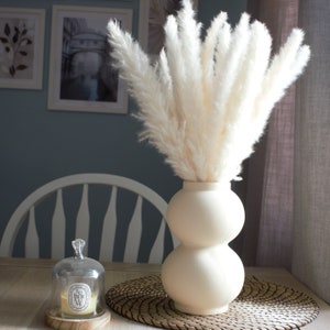 May include: A white ceramic vase with a rounded, bulbous shape holds a bouquet of dried pampas grass. The vase is sitting on a woven placemat.