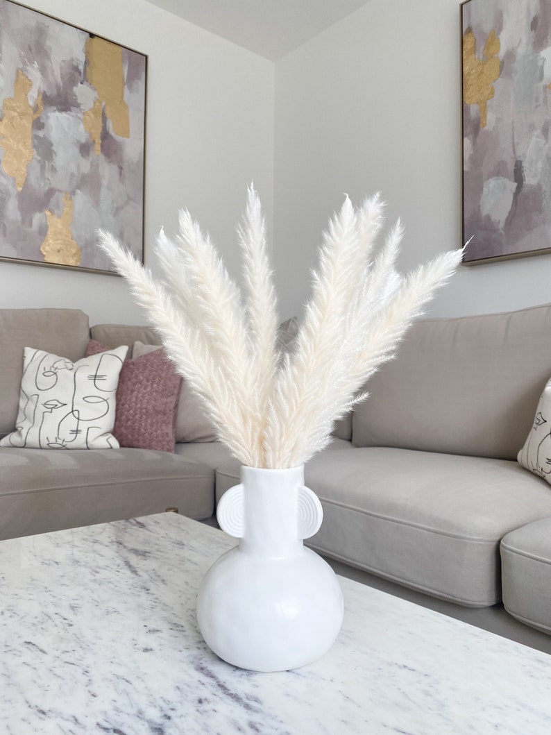 May include: White pampas grass in a white ceramic vase with two round handles. The vase is sitting on a white marble table in front of a light grey couch.
