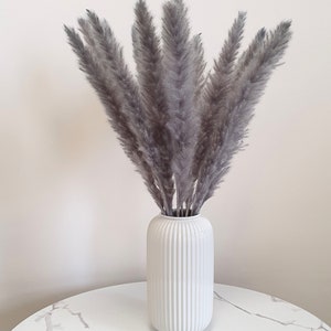 May include: A bouquet of gray pampas grass in a white ceramic vase with vertical ridges. The vase is sitting on a white marble table.
