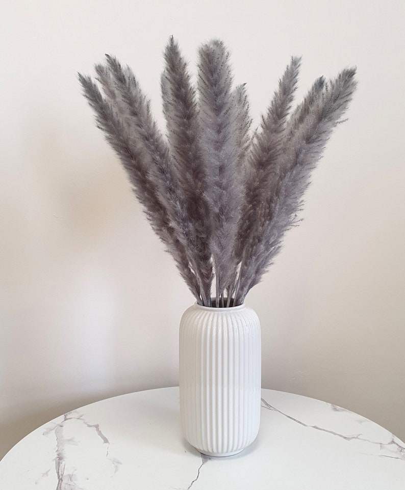 May include: A bouquet of gray pampas grass in a white ceramic vase with vertical ridges. The vase is sitting on a white marble table.