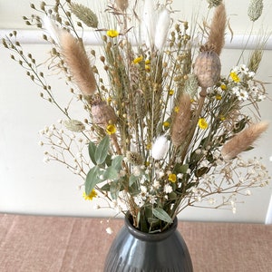 May include: A dried flower arrangement in a dark gray ceramic vase. The bouquet features a variety of dried grasses, pampas grass, and small white and yellow flowers.