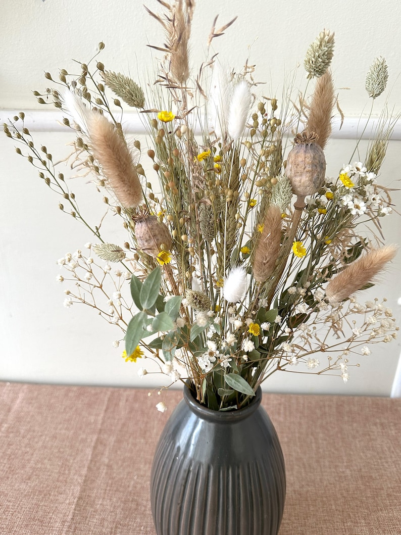 May include: A dried flower arrangement in a dark gray ceramic vase. The bouquet features a variety of dried grasses, pampas grass, and small white and yellow flowers.