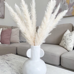 May include: White pampas grass in a white ceramic vase with two round handles. The vase has a round body and a narrow neck. The vase is sitting on a white marble table.