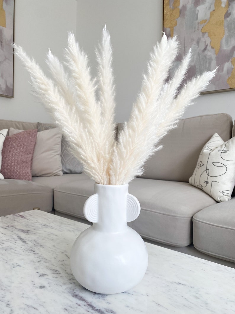 May include: White pampas grass in a white ceramic vase with two round handles. The vase has a round body and a narrow neck. The vase is sitting on a white marble table.