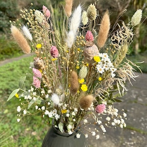 May include: A dried flower bouquet in a dark green vase. The bouquet includes white, brown, and beige dried grasses, pink and yellow wildflowers, and white baby's breath.