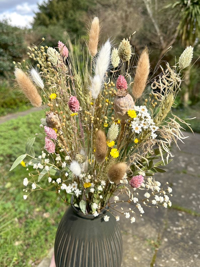 May include: A dried flower bouquet in a dark green vase. The bouquet includes white, brown, and beige dried grasses, pink and yellow wildflowers, and white baby's breath.