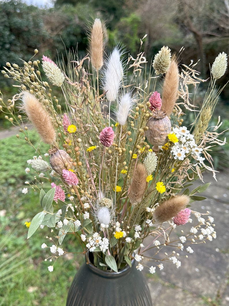 May include: A dried flower arrangement in a dark green vase. The bouquet features a variety of dried grasses, flowers, and seed pods in shades of white, pink, yellow, and brown.