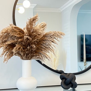 May include: A white ceramic vase with a round base holds a bouquet of dried pampas grass. The vase is on a black and glass table in front of a round black mirror.