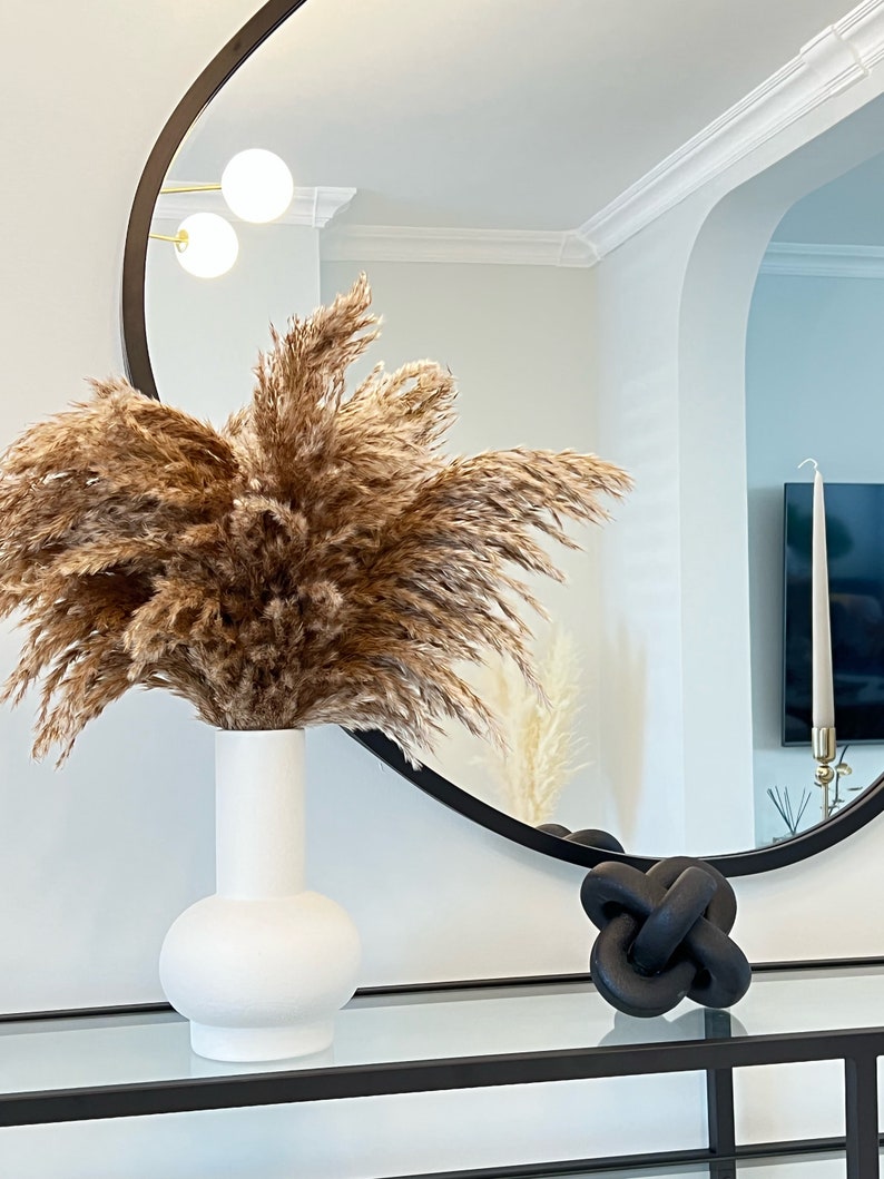 May include: A white ceramic vase with a round base holds a bouquet of dried pampas grass. The vase is on a black and glass table in front of a round black mirror.