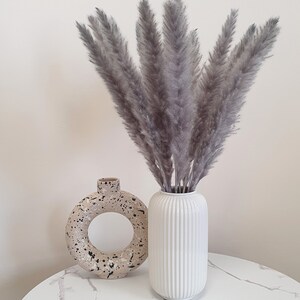 May include: A bouquet of dried pampas grass in a white vase with a textured surface. The vase is sitting on a white marble table next to a beige ceramic vase with black speckles.