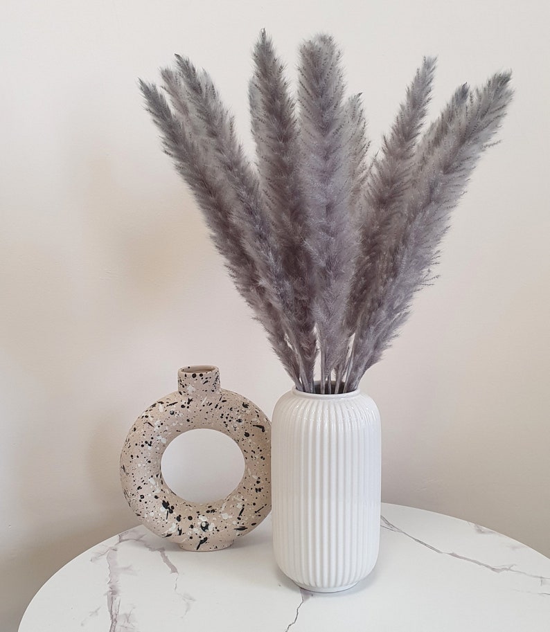 May include: A bouquet of dried pampas grass in a white vase with a textured surface. The vase is sitting on a white marble table next to a beige ceramic vase with black speckles.