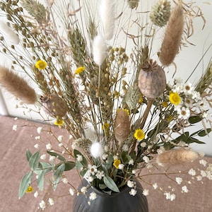 May include: A dried flower arrangement in a dark gray ceramic vase. The bouquet features a variety of dried grasses, white fluffy seed pods, and small yellow flowers.