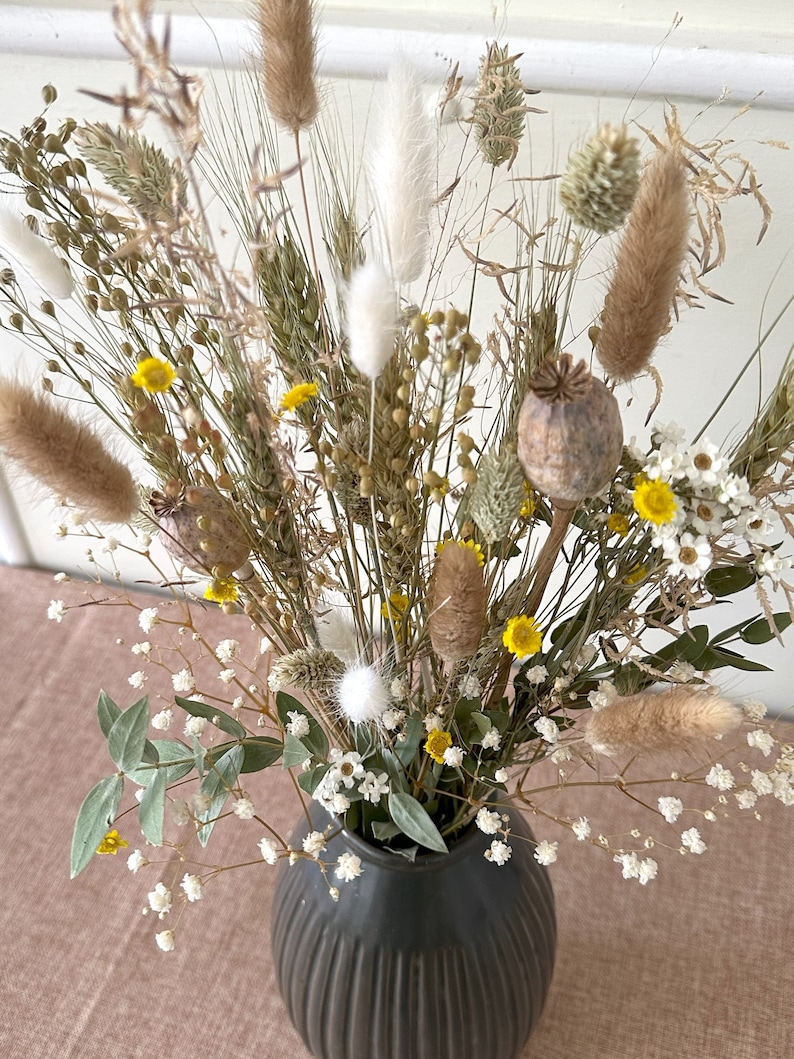 May include: A dried flower arrangement in a dark gray ceramic vase. The bouquet features a variety of dried grasses, white fluffy seed pods, and small yellow flowers.